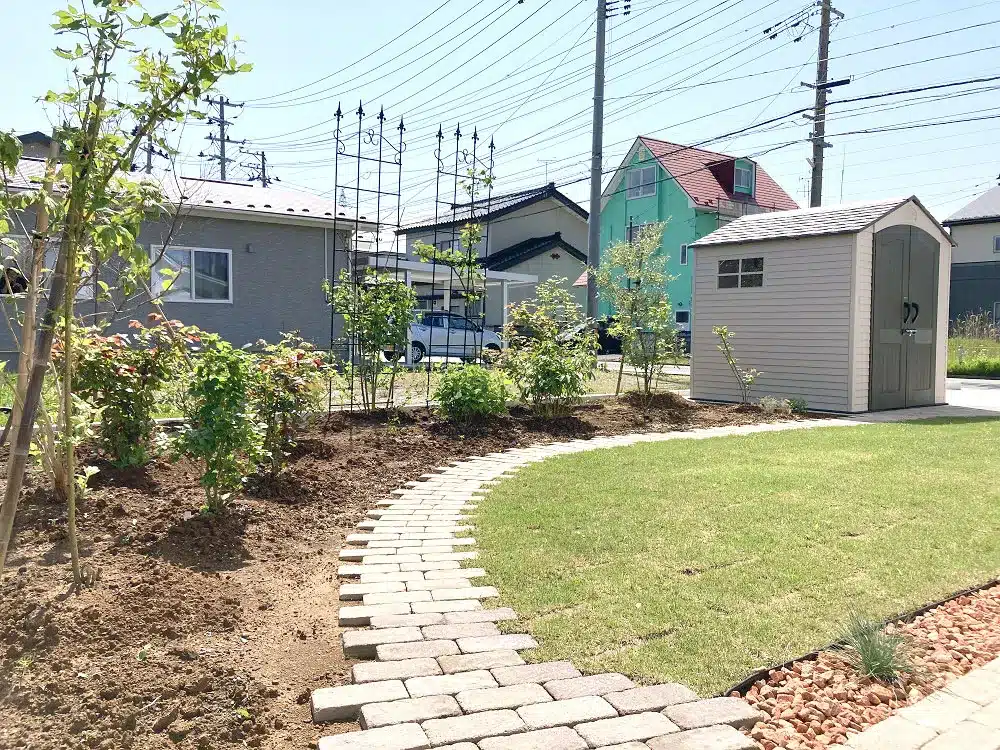 広々とした天然芝の庭に続くレンガ調の曲線的な園路と、庭の奥に立つデザイン物置。